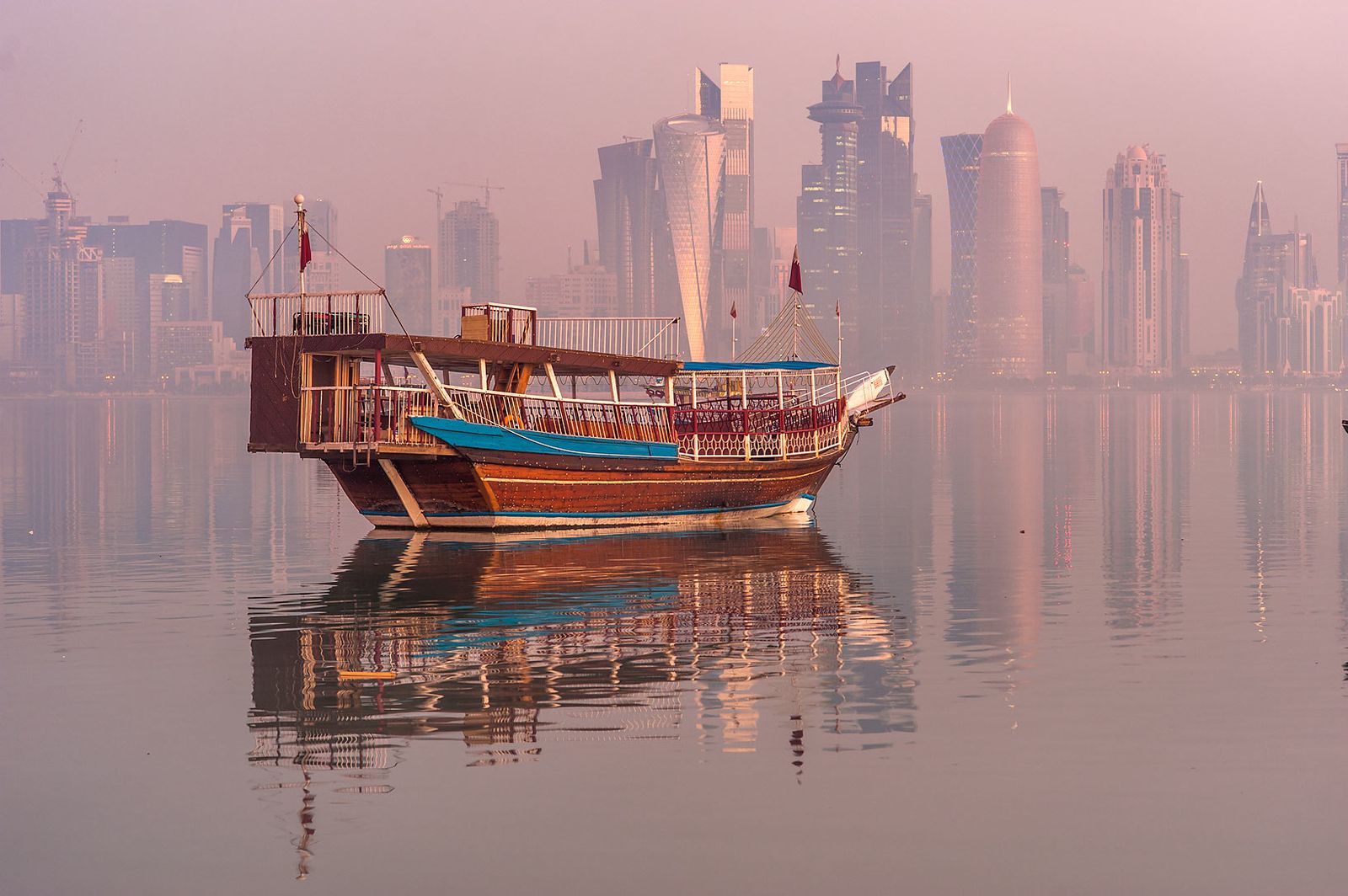 A wooden dhow reflected in still water off the Corniche, with West Bay towers fading into the haze